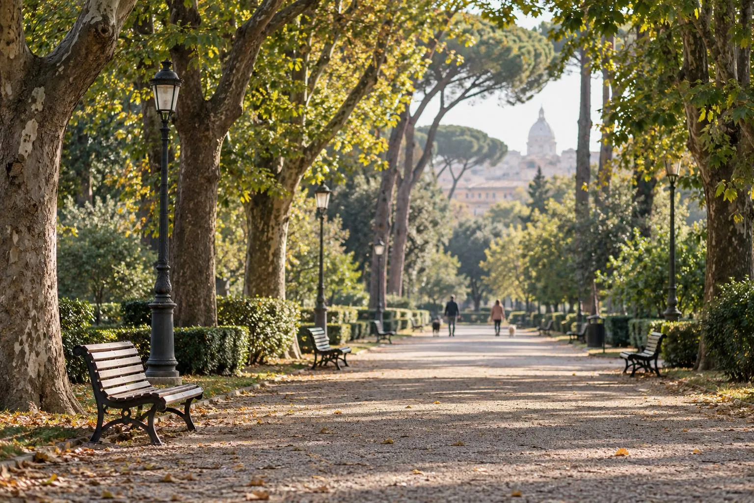 Viale alberato di un parco con panchine e lampioni, e due persone con i loro cani in lontananza